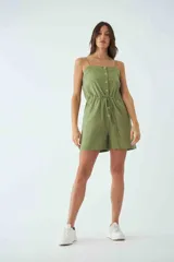 Monoprenda corta de color verde oliva, con tirantes finos ajustables, cierre frontal con botones y cintura elástica con cordón ajustable. Cuenta con bolsillos laterales.