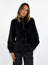 Camisa de pana negra con cuello alto, cierre frontal de botones y lazo ajustable en el cuello. Presenta mangas largas con puños abotonados y un corte holgado.