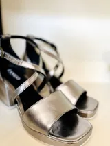 Sandalias plateadas de cuero metalizado, con taco cuadrado y plataforma. Presentan tiras cruzadas sobre el empeine y pulsera con hebilla al tobillo.