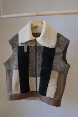 Chaleco sin mangas confeccionado con técnica de patchwork en cuero genuino y corderito, con diseño multicolor en tonos marrón, negro y blanco, solapas anchas y cierre frontal de cremallera.