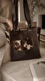 Bolso tipo tote bag de cuero marrón con detalles en pelo natural estampado tipo animal print de vaca, con asas largas para llevar al hombro y bolsillo exterior.