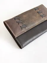 Billetera de cuero con diseño bicolor en tonos marrón y chocolate, presenta detalles de costuras decorativas en forma de cruces en la solapa frontal y cierre con broche.
