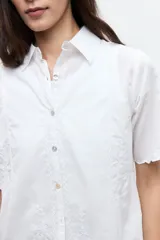 Camisa blanca de manga corta con sutiles detalles bordados tonales, cierre frontal de botones y cuello camisero.