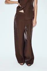 Pantalón de tiro alto color marrón con tejido de efecto brillo y pernera recta. Cuenta con bolsillos delanteros y cierre frontal con cremallera, botón interior y gancho.