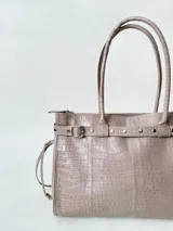 Bolso grande de cuero marrón con textura tipo cocodrilo, estructura rígida y asas para llevar al hombro. Tiene una tira con tachas y hebillas en cada extremo, también lleva lazos en los costados.