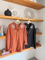 Camisa de color terracota con cuello camisero, cierre frontal de botones y mangas largas abullonadas. Presenta un delicado bordado floral en color blanco en la zona del pecho y los hombros, con un diseño de corte holgado.
