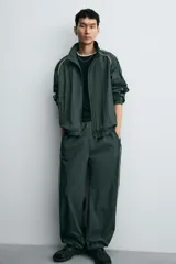 Pantalón jogger de corte relajado confeccionado en tejido técnico de color verde oscuro. Presenta cintura elástica con cordón interior, bolsillos laterales con cierre y bajo ajustable con elástico. Incluye detalles de líneas en contraste a lo largo de las piernas.