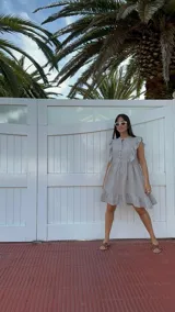 Vestido corto color gris claro, con escote redondo, mangas cortas con volados y falda con volados en el ruedo. Tiene botones en el frente.