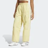 Pantalón deportivo amarillo pálido de corte holgado con cintura y puños elásticos. Presenta tres tiras blancas verticales a lo largo de cada pierna y logo de Adidas bordado en blanco en la cadera izquierda.