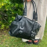 Bolso tipo shopper de eco cuero negro con detalles de correas frontales y herrajes metálicos plateados. Cuenta con doble asa de hombro y cierre superior.