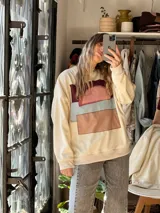 Buzo de friza color natural de calce relajado, con cuello redondo, mangas largas y puños elastizados. Presenta una composición abstracta de parches textiles cosidos a mano en la parte frontal en tonos bordó, rosado, celeste y tostado.