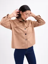 Chaqueta de gamuza sintética color beige, de corte recto, con cuello camisero, bolsillos frontales con solapa y cierre de botones oscuros.