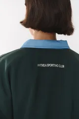 Buzo estilo rugby de corte oversized, color verde oscuro, con cuello tipo camisa en denim azul claro y cierre frontal parcial. Presenta un recorte horizontal en el pecho, logo bordado y tiras laterales en las mangas en contraste.