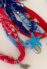 Collar tipo gargantilla confeccionado con un pañuelo bandana azul con estampado blanco. Presenta un dije central en forma de estrella de mar color turquesa, un dije metálico pequeño en forma de concha y cuentas de colores rojo y turquesa.