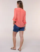 Blusa de calce relajado color salmón, confeccionada en tela liviana con textura sutil. Presenta escote en V con abotonadura frontal, bolsillo parche en el pecho, mangas largas regulables con presilla y ruedo con opción de anudar en el frente.