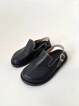 Zueco de cuero negro con textura granulada, punta cerrada y pespuntes blancos contrastantes. Posee una tira trasera ajustable con hebilla metálica plateada y suela de goma negra.