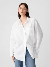 Camisa de corte oversize confeccionada en popelina de algodón, color blanco. Presenta cuello clásico, cierre frontal con botones, mangas largas con puños abotonados y un bolsillo aplicado en el pecho.