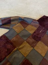 Buzo de cuero genuino con diseño de patchwork en tonos marrón, gris y bordó. Presenta cuello alto con cierre frontal parcial, bolsillo frontal tipo canguro y detalles de piel sintética en cuello, cierre y bolsillo.