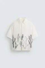 Camisa de calce relajado confeccionada en tejido ligero de lino y viscosa, con cuello solapa, manga corta y cierre frontal de botones. Presenta un diseño estampado de cactus en color gris en la parte inferior sobre un fondo blanco roto.