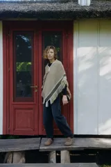 Poncho artesanal de lana en color beige, con diseño de tejido rústico y detalles de guardas geométricas en negro y blanco en los bordes. Presenta terminaciones con flecos largos en todo el contorno.