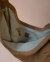 Bolso tipo hobo de gran tamaño, confeccionado en gamuza color marrón oscuro. Presenta un diseño artesanal con costuras visibles y detalles de tientos en el cuerpo, asa integrada con pespuntes a mano y cierre superior.