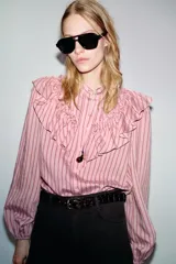 Camisa de manga larga con cuello subido, cierre frontal con botones y detalle de volantes en el pecho. Presenta un diseño a rayas verticales y puños elásticos.