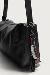 Bolso tipo bandolera de color negro, con compartimento principal con cierre de cremallera y bolsillo interior. Presenta un asa de hombro ajustable y un detalle decorativo de charms colgantes en uno de los laterales.
