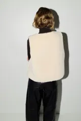 Chaleco de efecto peludito color beige, con cuello redondo, cierre frontal de botones y ribetes en contraste marrón oscuro. Incluye bolsillos laterales discretos y forro interior.