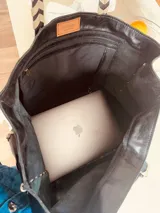 Bolso tipo tote de cuero negro con bolsillo externo y llavero interno. Tiene manijas cortas con diseño chevron y costuras blancas a contraste.