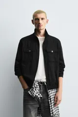 Cazadora de hombre color verde oliva, estilo bomber o Harrington, con cuello subido, cierre frontal de cremallera y puños y bajo elásticos. Presenta bolsillos de plastrón con solapa en el pecho y bolsillos de vivo en la cadera.