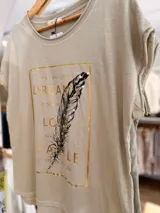 Remera de algodón color beige con corte relajado y diseño asimétrico, más larga en la parte posterior. Presenta un estampado frontal rectangular con letras doradas, una pluma ilustrada y detalles de pedrería decorativa.