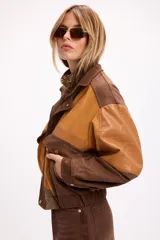 Chaqueta bomber de diseño combinado en tonos tierra, confeccionada con paneles de gamuza y detalles en cuero sintético. Presenta cierre frontal con broches metálicos, bolsillos frontales con tapa y puños elásticos.