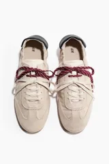 Calzado deportivo de gamuza color beige, con cordones intercambiables en tonos blanco y bordó, detalle de talón plegable con refuerzo negro y suela plana de goma acanalada.