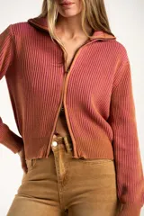 Campera tejida de algodón con diseño acanalado, cuello alto y cierre frontal completo. Presenta un patrón bicolor en tonos terracota y rosado, con puños y cintura elásticos.