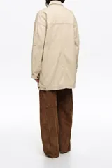 Chaquetón de corte recto color beige, confeccionado en algodón, con cierre frontal de cremallera oculta y botones a presión. Posee cuello camisero, mangas largas, bolsillos con solapa en el pecho, bolsillos tipo plastrón en la parte inferior y un pequeño bolsillo con cierre en la manga.