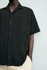 Camisa de corte relajado en color negro, confeccionada en tejido de algodón. Presenta cuello tipo bowling, manga corta y cierre frontal con botones. Cuenta con bordados lineales de hojas en la parte delantera.