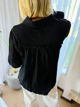 Camisa de manga larga confeccionada en muselina negra, con cuello clásico y cierre frontal de botones. Presenta un delicado bordado floral a tono sobre el hombro y bolsillo frontal.