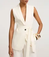 Chaleco sastre alargado color blanco hueso con finas rayas verticales. Presenta cuello tipo blazer, cierre cruzado con un botón metalizado y un lazo ancho para ajustar a la cintura.
