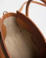 Cartera mediana color tostado, con doble asa de mano, detalles de herrajes dorados y correa larga regulable y desmontable. Cuenta con placa metálica frontal con el logo de la marca.