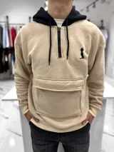Canguro oversize beige con capucha y cordones negros, cierre frontal parcial con cremallera, bolsillo canguro delantero con solapa y pequeño bordado negro en el pecho.