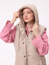 Chaleco largo acolchado color beige, con capucha forrada en sherpa, cierre frontal con cremallera y botones a presión, y bolsillos laterales.