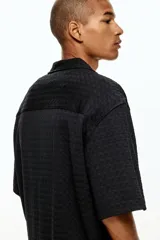 Camisa negra de manga corta con cuello resort, tapeta lisa y bajo recto con aberturas laterales. Corte holgado.