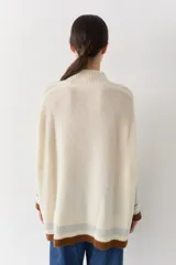 Buzo tejido de punto con cuello alto, diseño holgado y aberturas laterales. Presenta un cuerpo en color azul melange con detalles de franjas en color crema y verde en los puños y el ruedo. Incluye un pequeño bordado en el pecho.