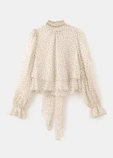 Blusa de gasa color beige con estampado de lunares negros, cuello alto con lazo integrado y mangas largas con puños amplios.