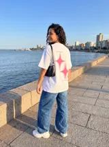 Remera oversize negra con estampado trasero en color rosa vibrante. El estampado incluye dos figuras de estrella con efecto de brillo y texto en cursiva que dice "Limits are optional", "Rush the moment" y "Own the night".