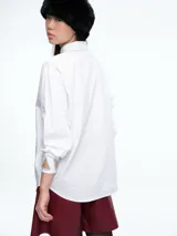 Camisa blanca de corte holgado con cuello clásico y cierre frontal de botones. Presenta un diseño distintivo en los puños con aberturas decoradas con pedrería brillante.