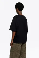 Camiseta negra de corte recto y largo medio, confeccionada en algodón orgánico. Presenta escote redondo, mangas cortas amplias con costuras raglán y detalles de cortes diagonales en el cuerpo.
