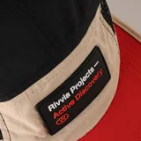 Gorro de running tipo jockey con paneles de malla negra, panel frontal beige con parche rectangular negro que dice "Rivvia Projects - Active Discovery" y visera roja.