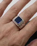 Anillo de acero tipo sello con piedra central rectangular color azul y diseño lateral de flor de lis.