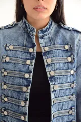 Chaqueta de denim estilo militar con diseño estructurado, cuello mao y cierre frontal con múltiples botones metálicos decorativos dispuestos en filas horizontales.